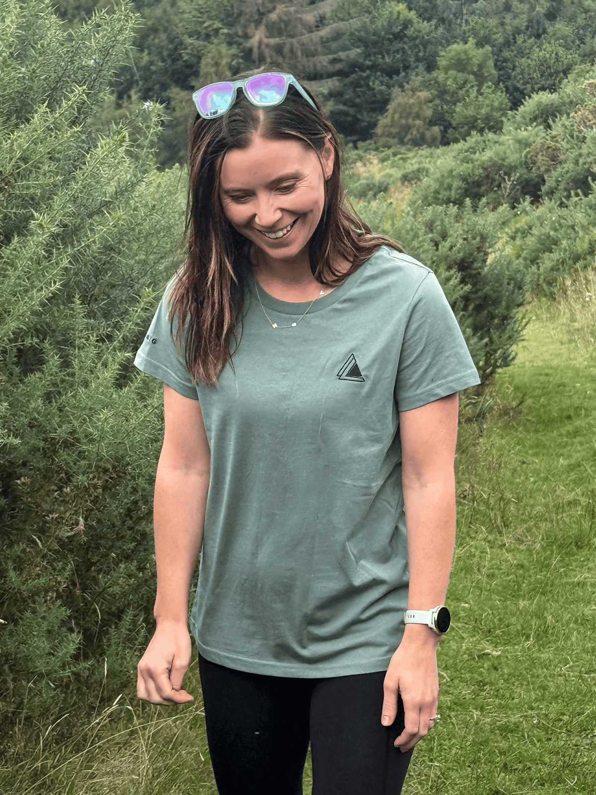 Smiling woman wearing the Stravaig Womens Everyday Trail T-Shirt in Machair Sage on a woodland path
