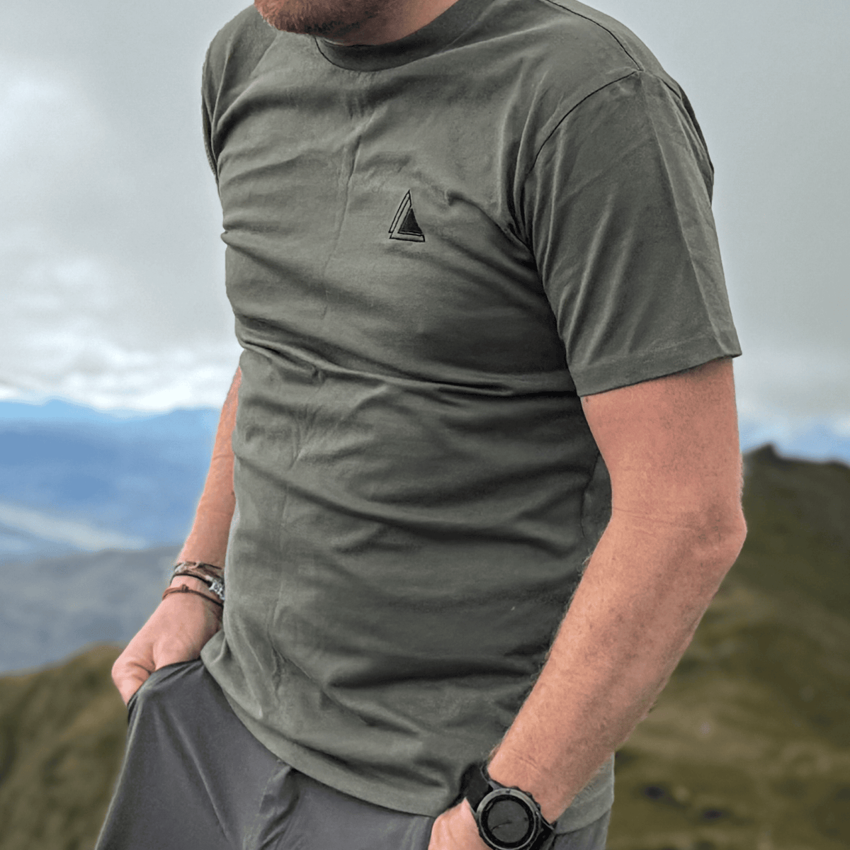 Man in the Highland Moss Stravaig t-shirt on a hilltop with mountain views behind