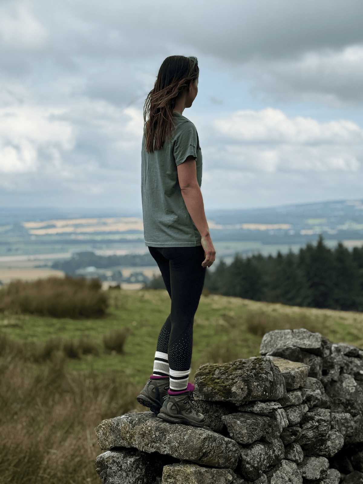 Woman in a Machair Sage Stravaig t-shirt standing on a dry stone wall looking out over moorland