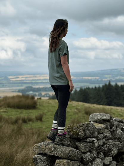 Woman in a Machair Sage Stravaig t-shirt standing on a dry stone wall looking out over moorland