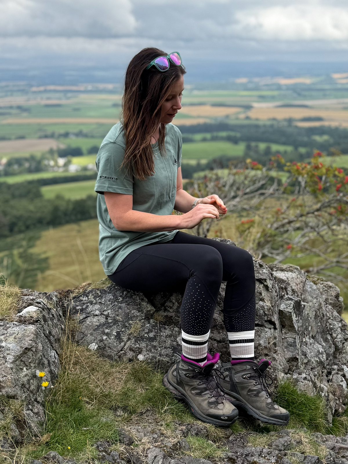 Woman in the Machair Sage Everyday Trail T-Shirt and hiking boots sitting on a rocky outcrop above the Scottish countryside