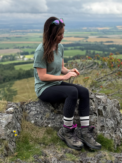Woman in the Machair Sage Everyday Trail T-Shirt and hiking boots sitting on a rocky outcrop above the Scottish countryside