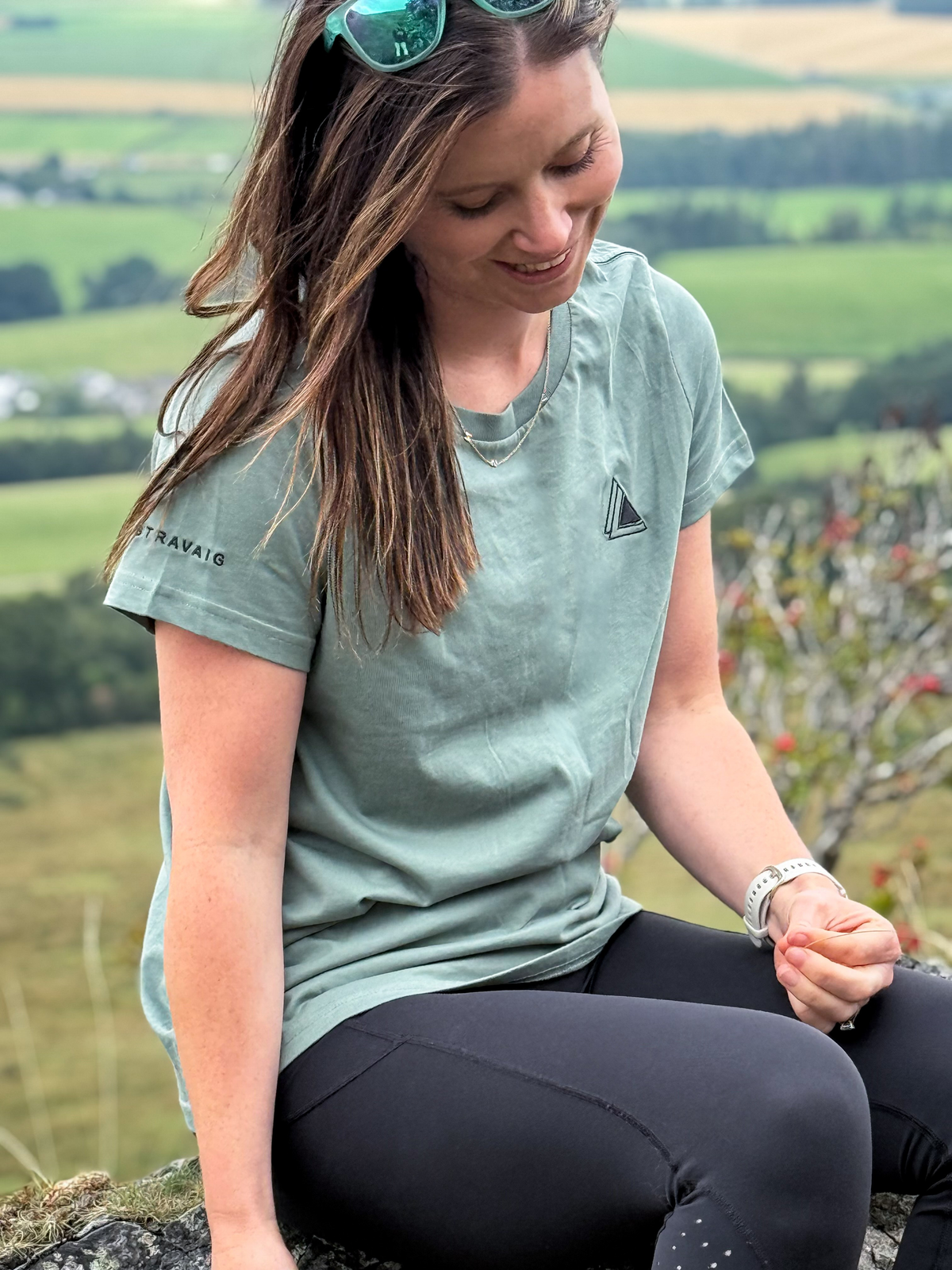 Woman in a Machair Sage Stravaig t-shirt sitting on a hilltop overlooking green fields