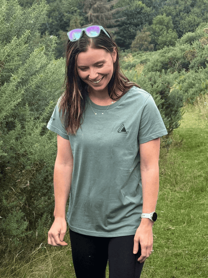 Smiling woman wearing the Stravaig Womens Everyday Trail T-Shirt in Machair Sage on a woodland path