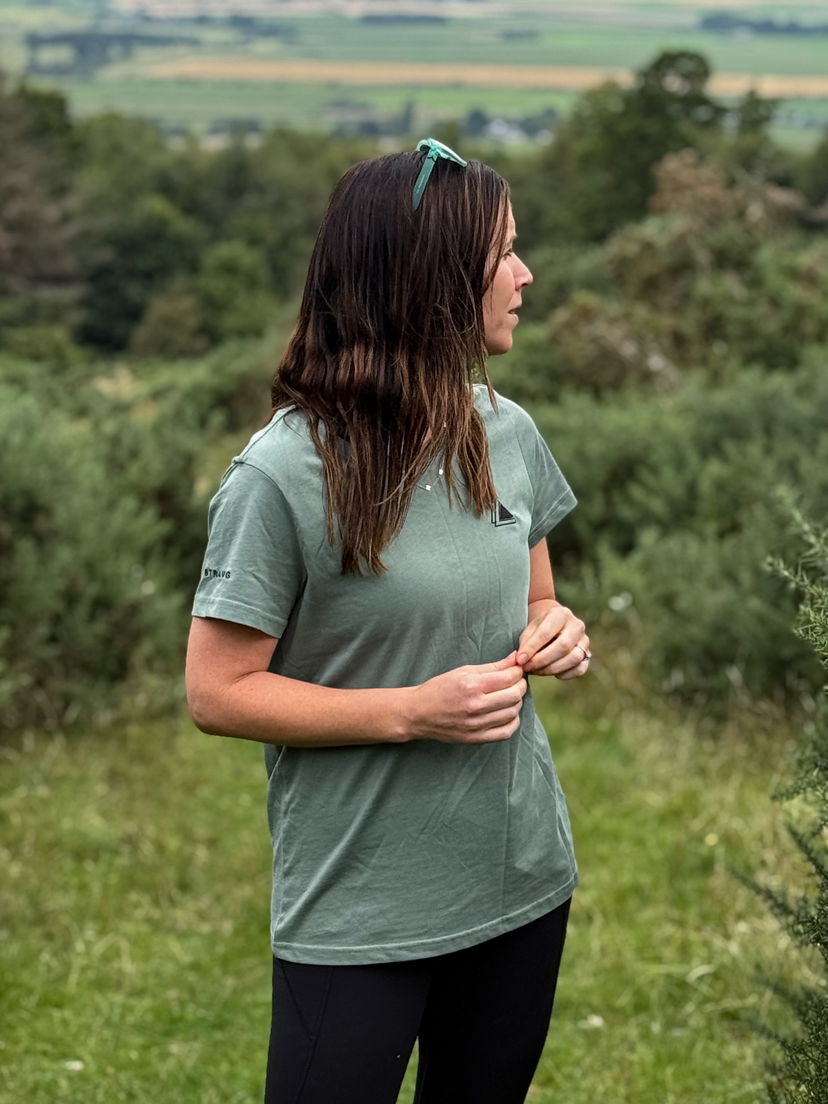 Side view of a woman in a Machair Sage Stravaig t-shirt standing among gorse on a hillside