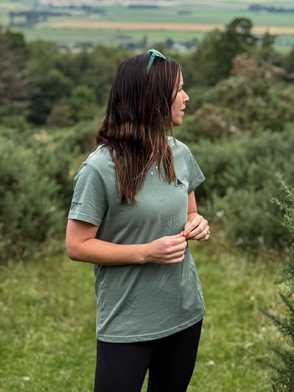 Side view of a woman in a Machair Sage Stravaig t-shirt standing among gorse on a hillside