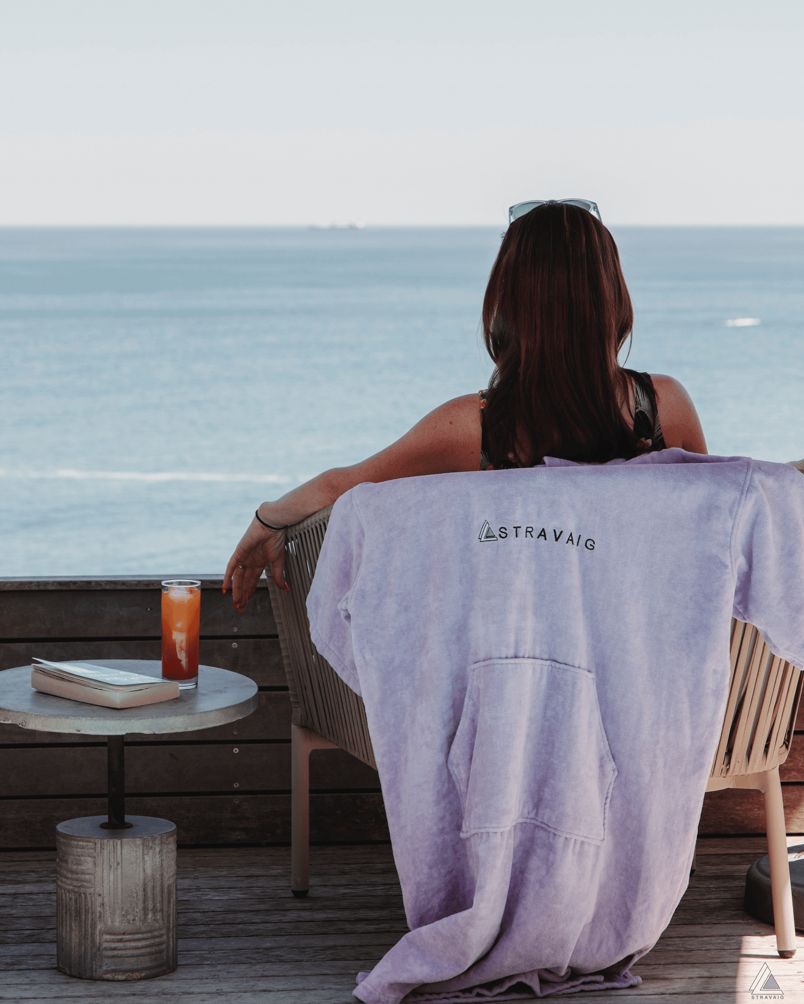 Dark haired woman sitting by the ocean with a Stravaig towelling robe draped over her chair, enjoying a drink.