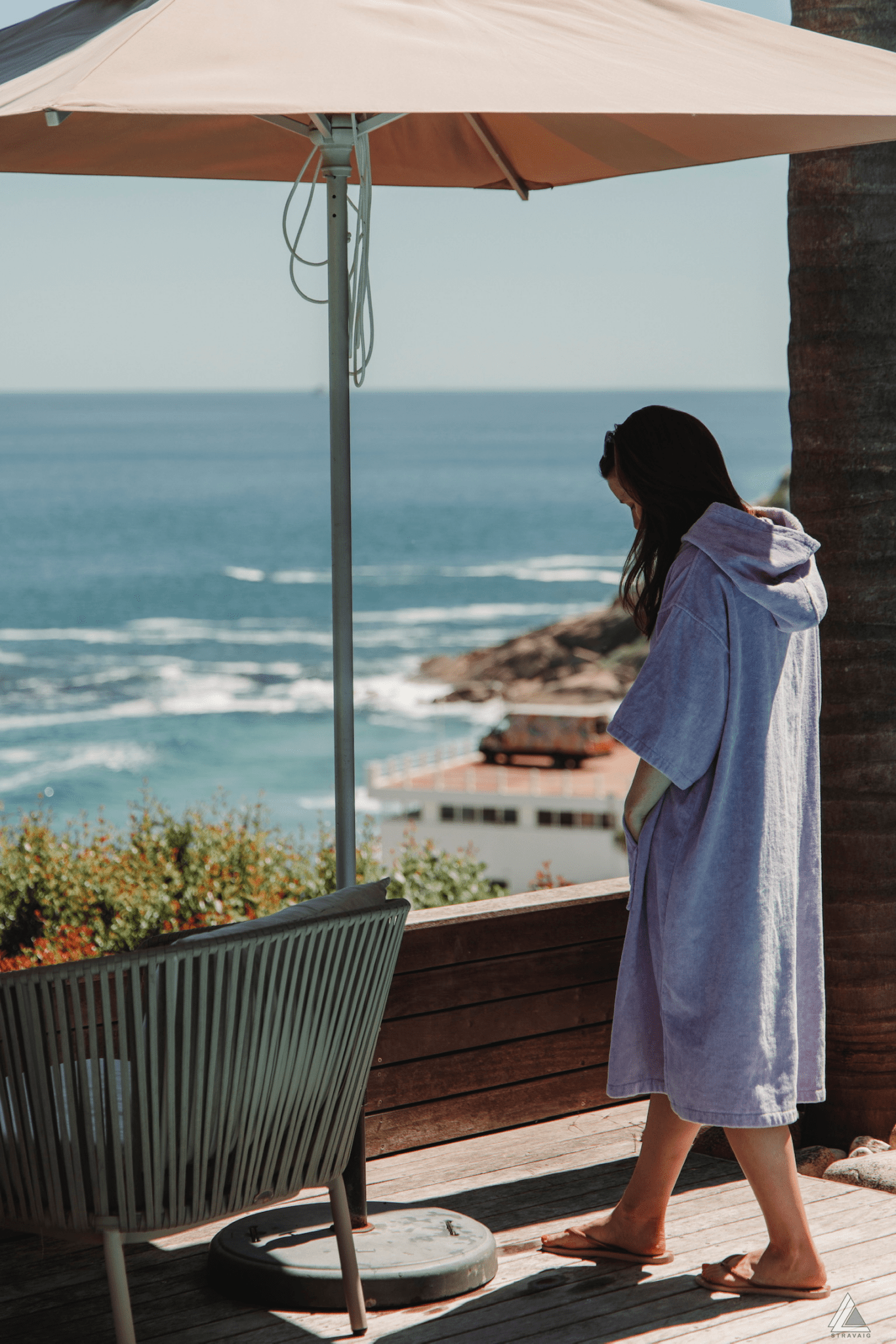 Woman in a Lilac Stravaig Adult Poncho Towel standing on a deck overlooking the ocean with a palm tree and chair in the foreground.