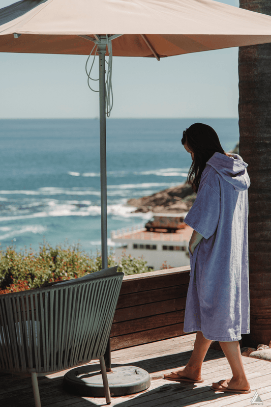 Woman in a Lilac Stravaig Adult Poncho Towel standing on a deck overlooking the ocean with a palm tree and chair in the foreground.