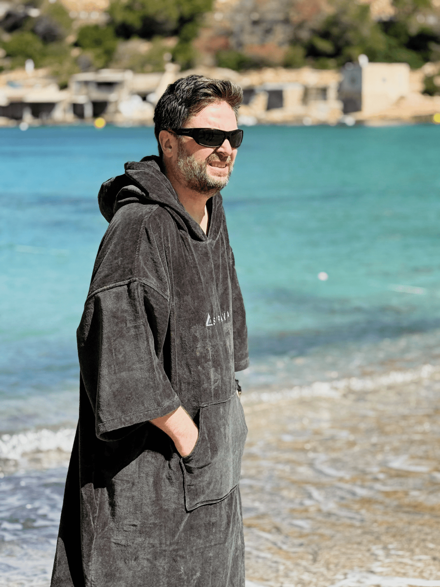 Man wearing a Sgàil Grey adult towel poncho on a beach with turquoise water and greenery in the background