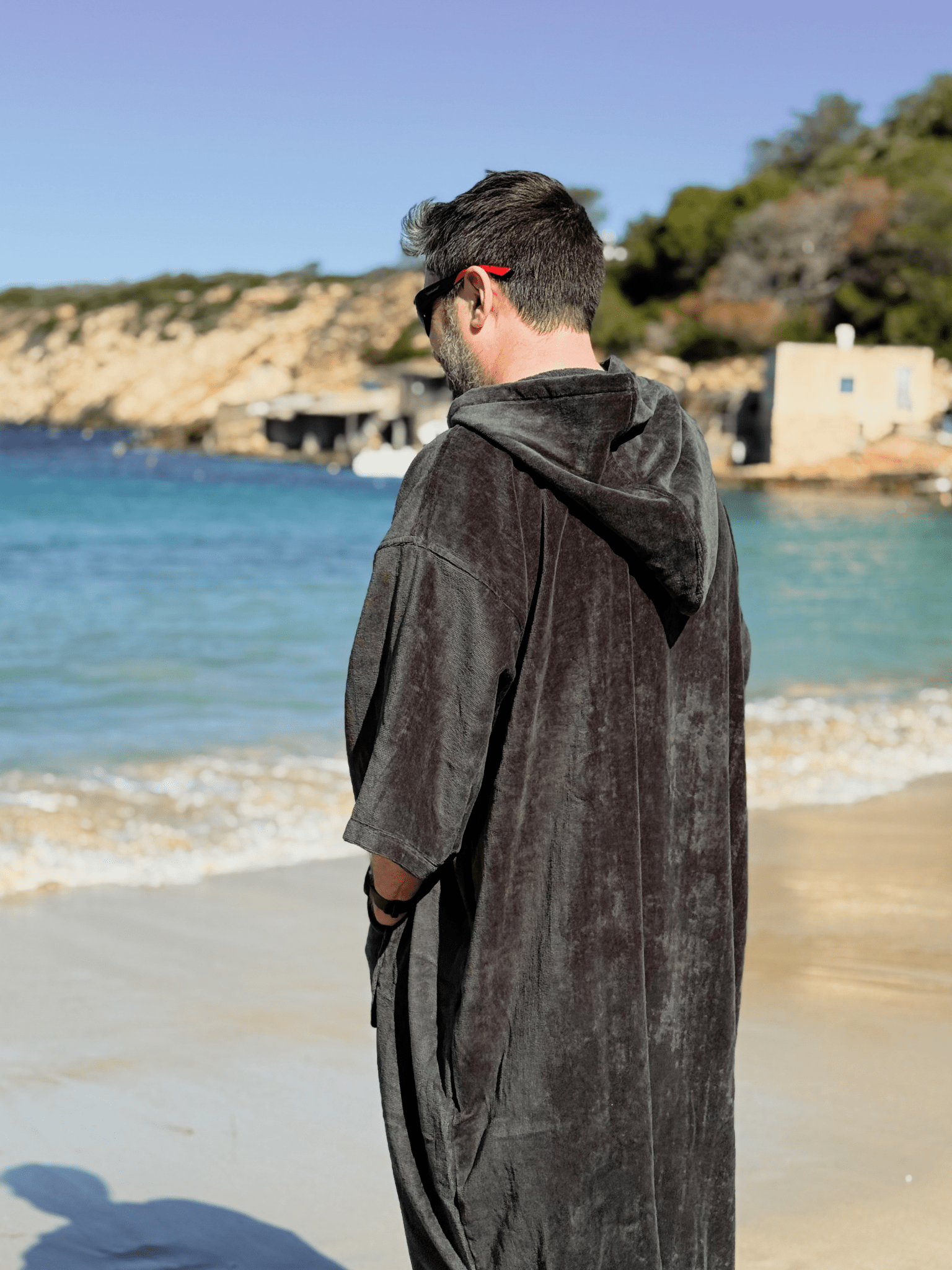 Man wearing a Sgàil Grey adult towel robe standing on a beach with ocean and cliffs in the background