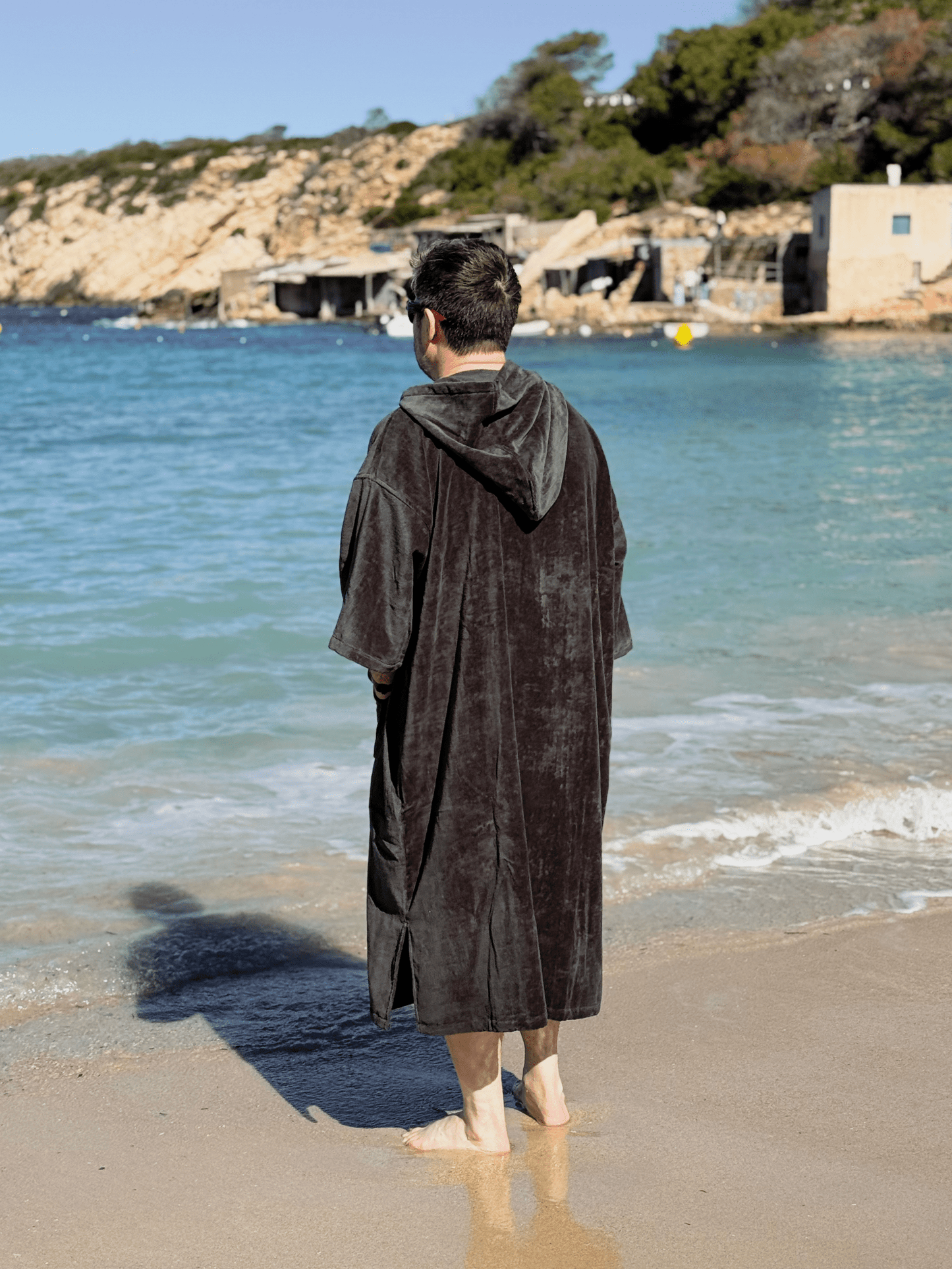Man wearing a Stravaig Sgàil Grey Adult Hooded Towel Poncho Changing Robe standing on a beach with ocean and rocky cliffs in the background