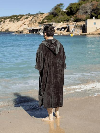 Man wearing a Stravaig Sgàil Grey Adult Hooded Towel Poncho Changing Robe standing on a beach with ocean and rocky cliffs in the background