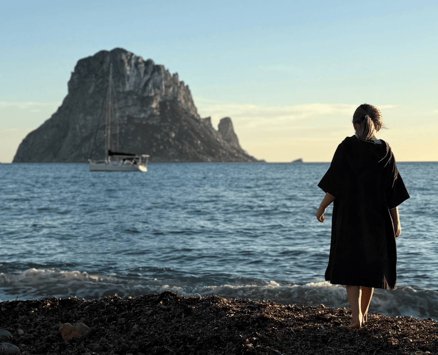 Child wearing a Stravaig Sgàil Grey kids changing robe  standing on a rocky beach with a large island and boat in the background.