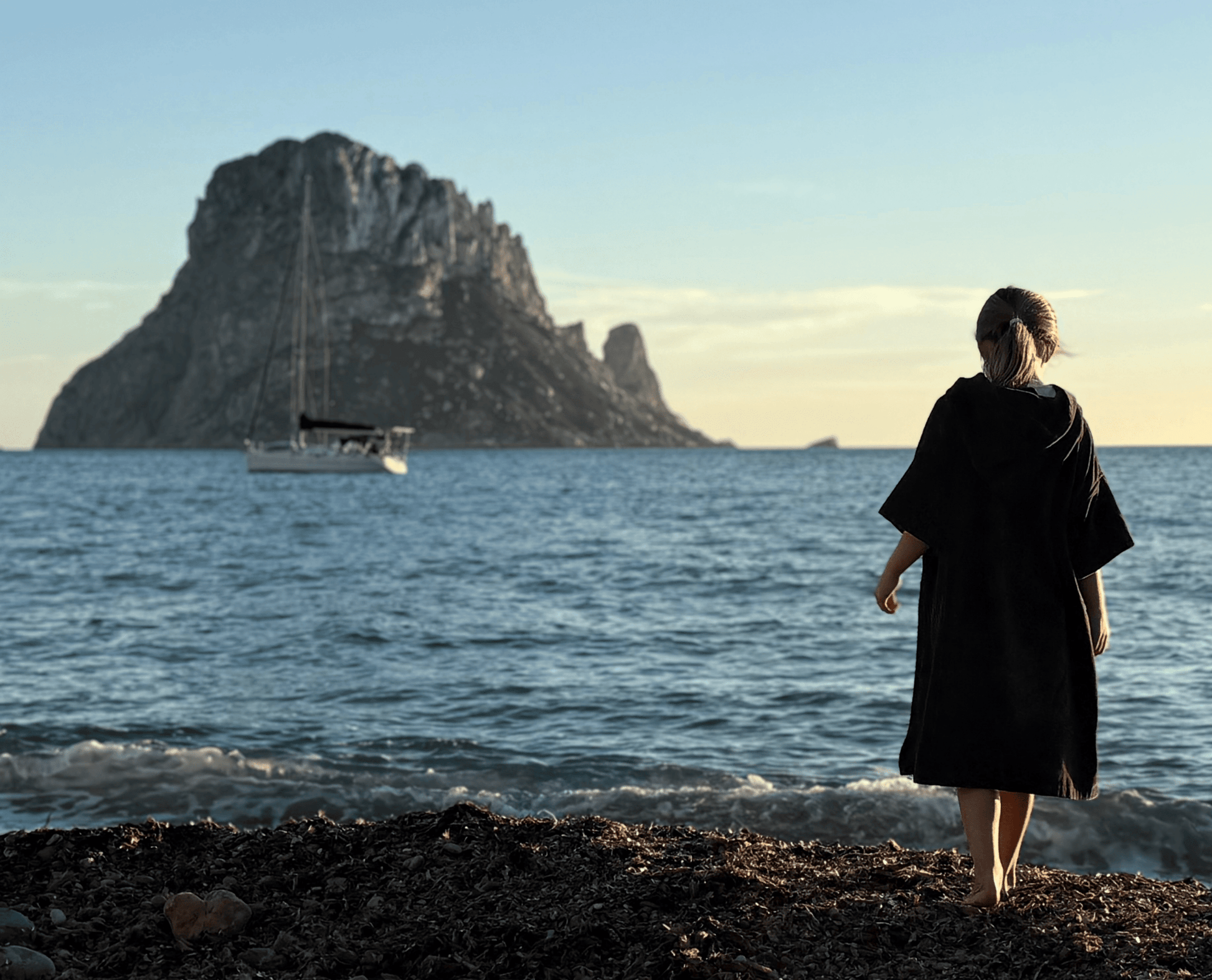 Child wearing a Stravaig Sgàil Grey kids changing robe  standing on a rocky beach with a large island and boat in the background.