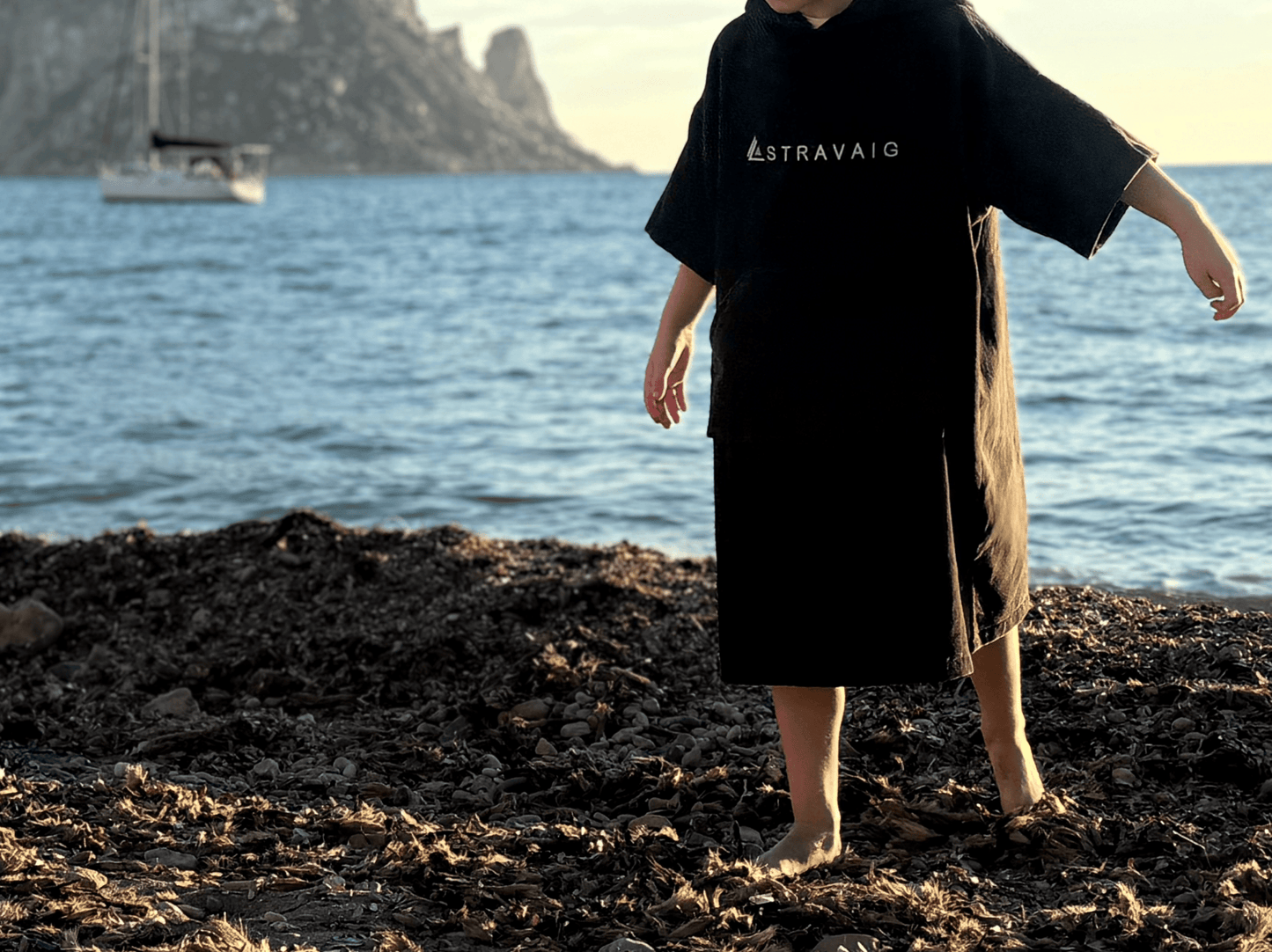 Child wearing a Stravaig Sgàil Grey hooded towel standing on a beach with water and mountains in the background.