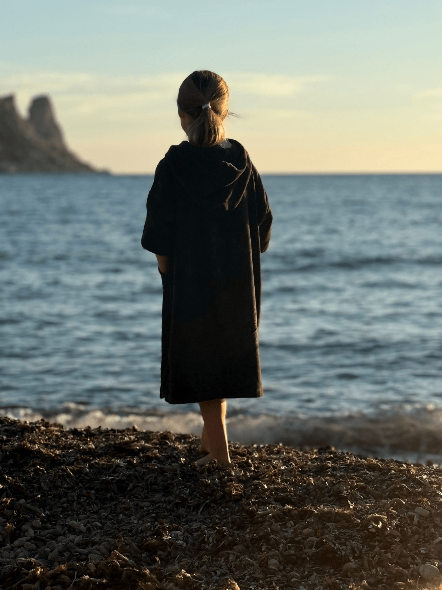 Child wearing a Stravaig Sgàil Grey childrens towelling robe standing on a beach looking out at the ocean during sunset.