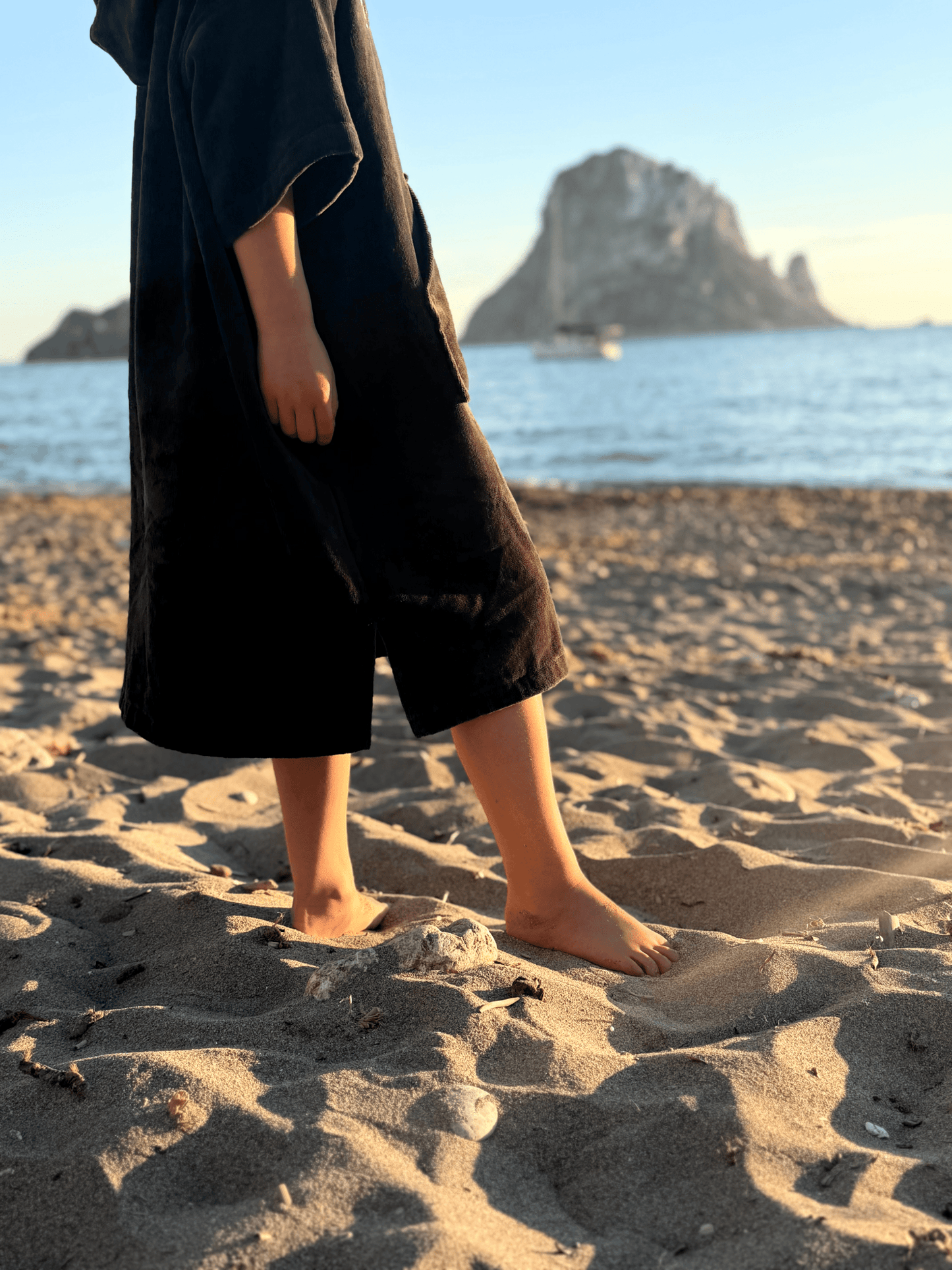 Child wearing a Stravaig Sgàil Grey kids towel robe standing on a sandy beach with a scenic background