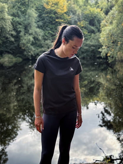 Woman wearing the Stravaig Womens Everyday Trail T-Shirt in Sgàil Grey beside a still woodland loch