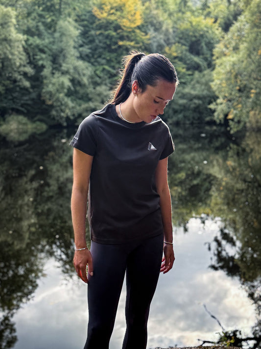 Woman wearing the Stravaig Womens Everyday Trail T-Shirt in Sgàil Grey beside a still woodland loch
