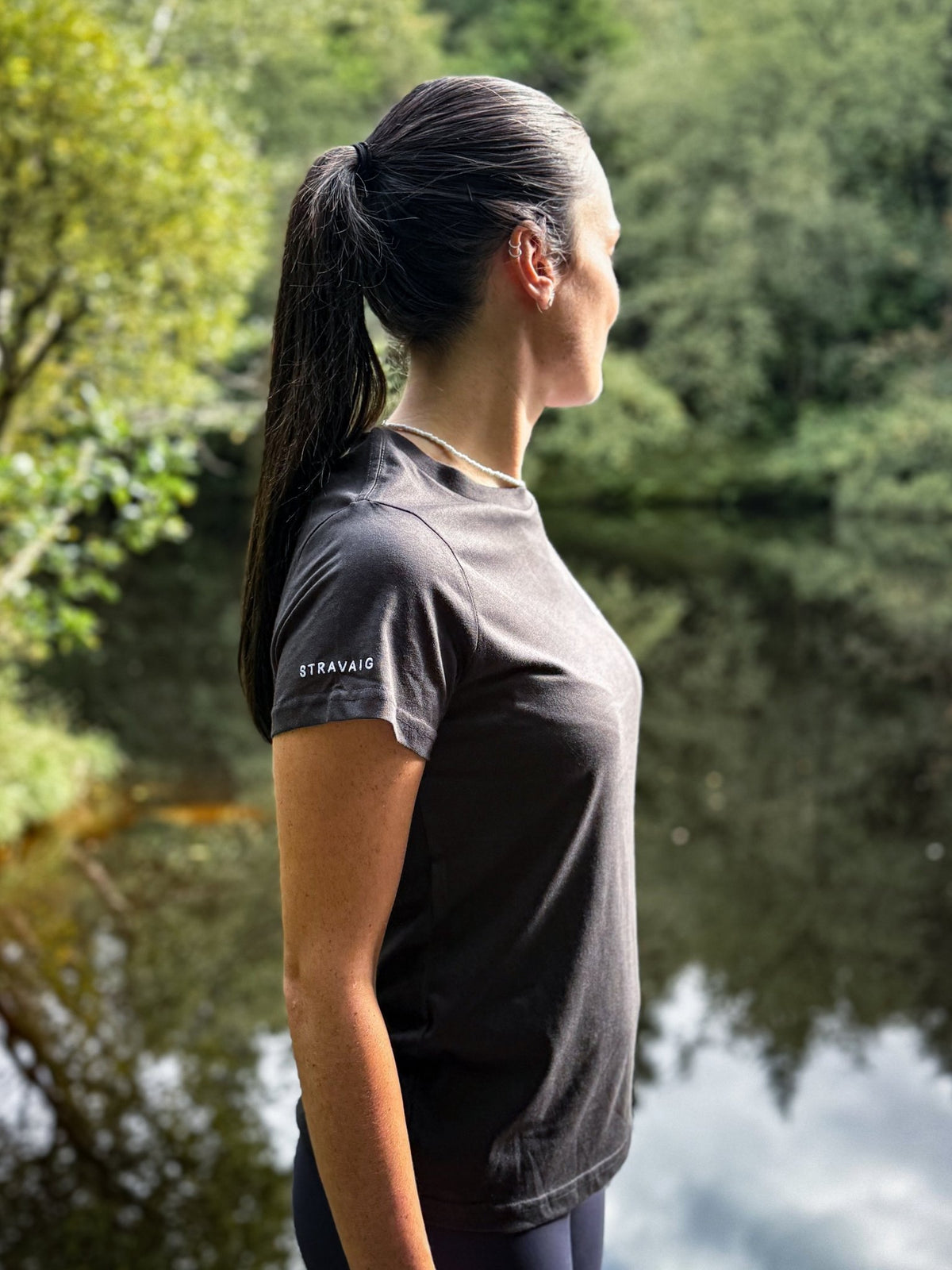 Profile view of a woman in a Sgàil Grey Stravaig t-shirt by a tree-lined loch
