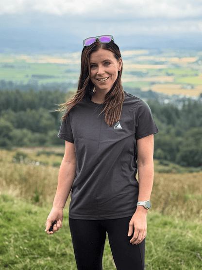 Woman in a Sgàil Grey Stravaig t-shirt on a grassy hilltop with farmland views behind